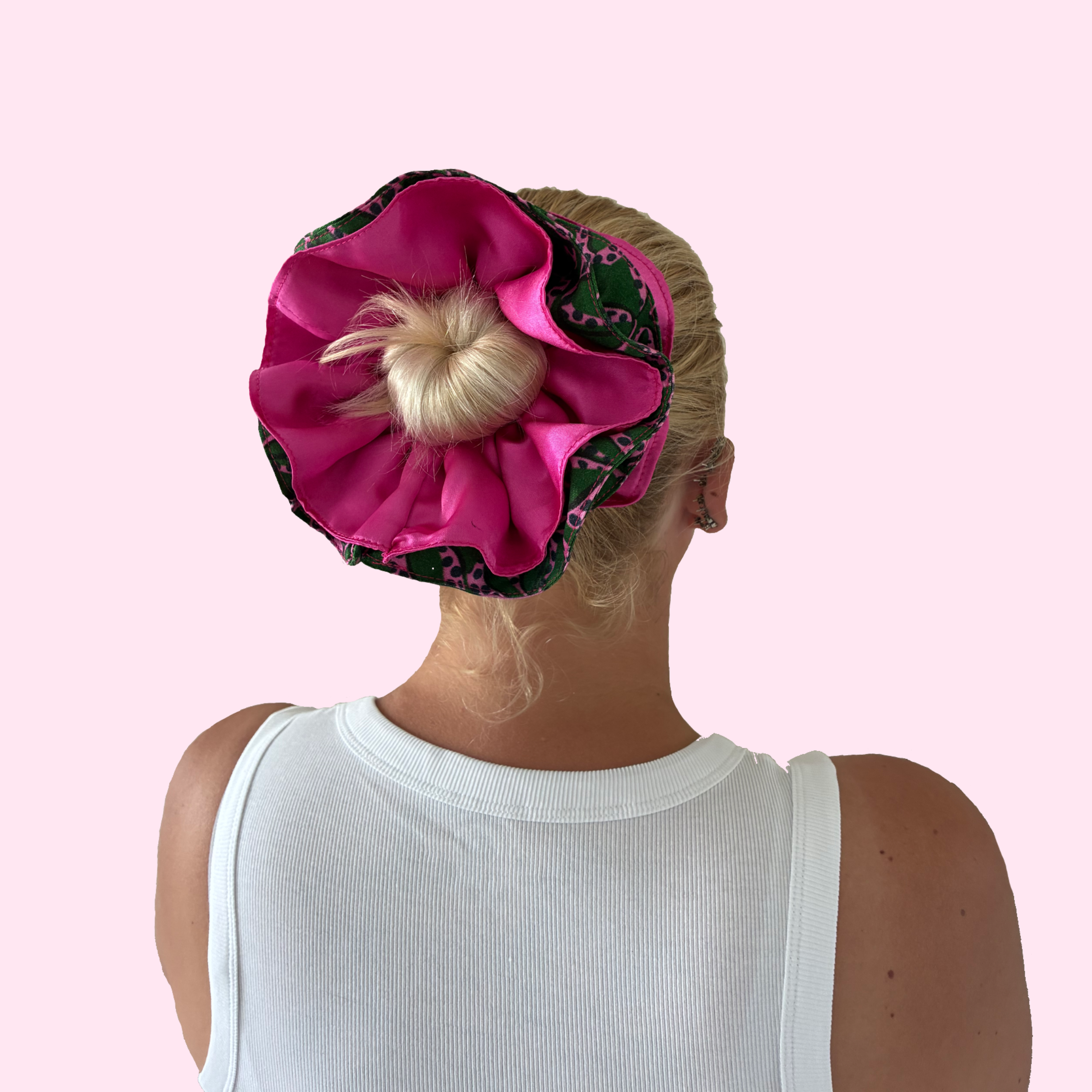 Person wearing a pink satin hair bun with pink bow print and green background. Reversible hair scrunchie, two-in-one hair accessory. Flip hair scrunchie inside out to reveal other colour and pattern 